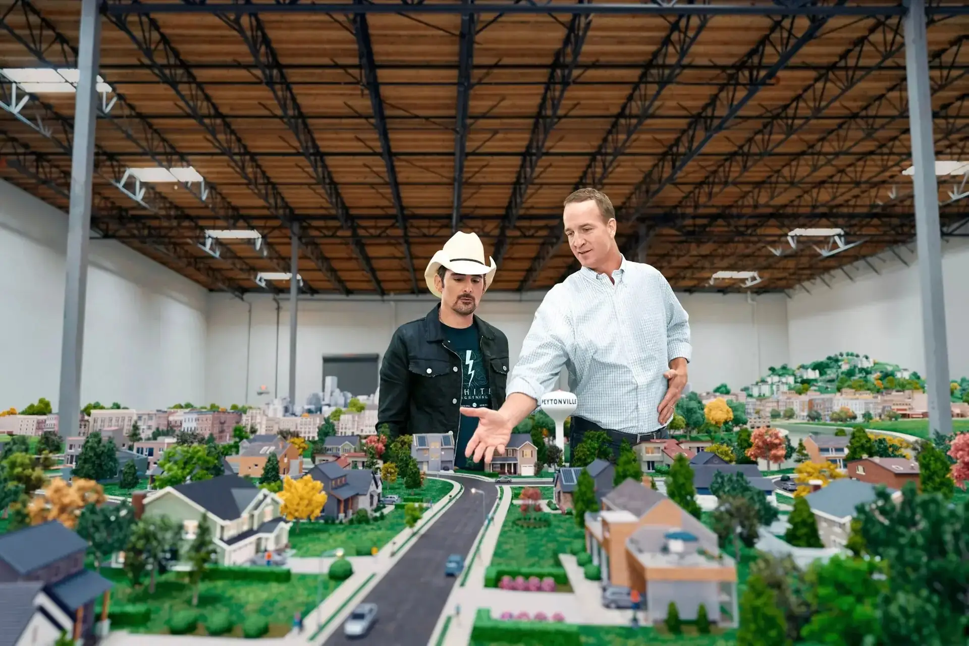 Two men stand behind a detailed model town layout, with a water tower that reads PEYTONVILLE.