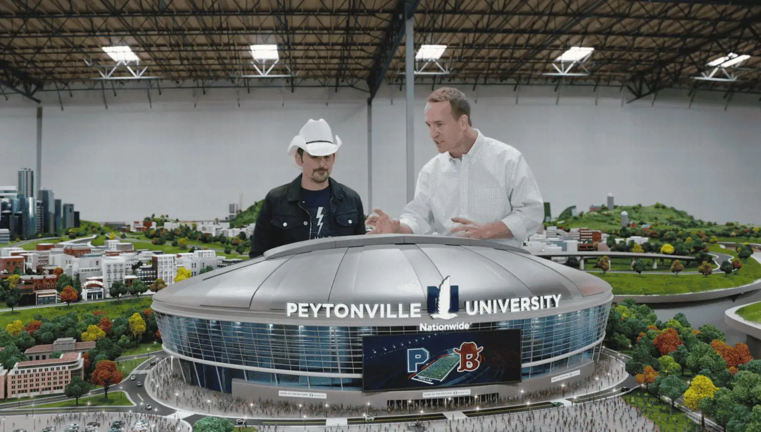 Two men stand behind a model stadium labeled Peytonville University.