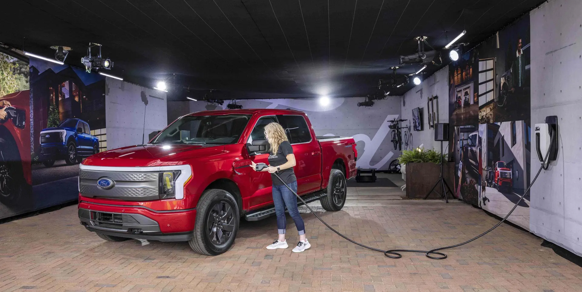 A woman charges a red Ford pickup truck plugged into a wall charger indoors.