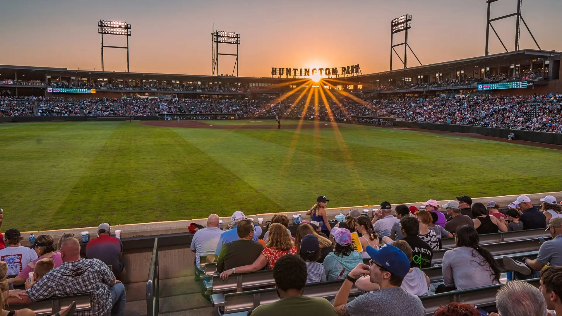 A baseball stadium during sunset, featuring Huntington Park signage above the stands filled with cheering fans and a large…